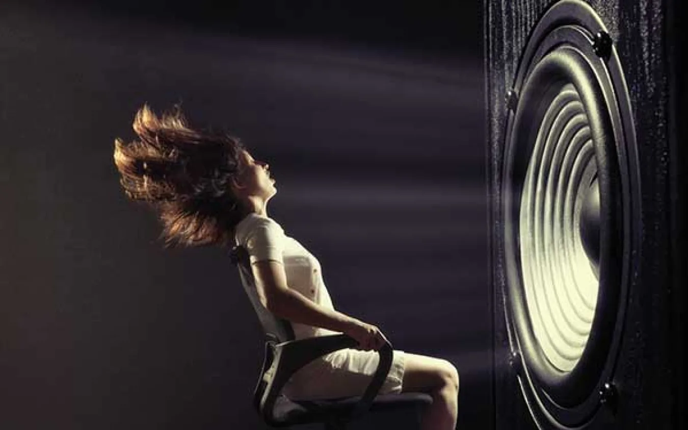A person seated against a large speaker, hair blown back by the airflow, testing the noise-to-energy concept